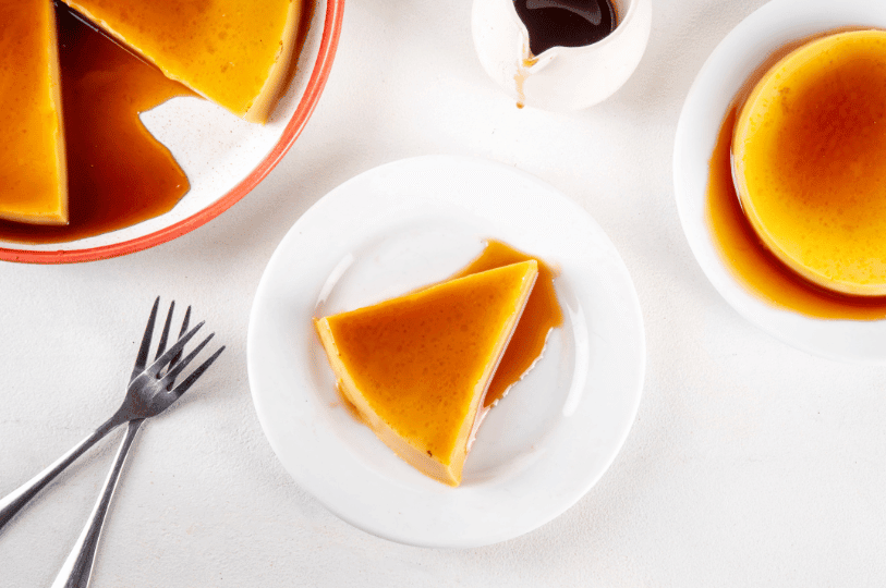Overhead view of a dessert spread featuring a sliced whole flan, a single triangular wedge on a plate, and a small pitcher of extra caramel sauce.