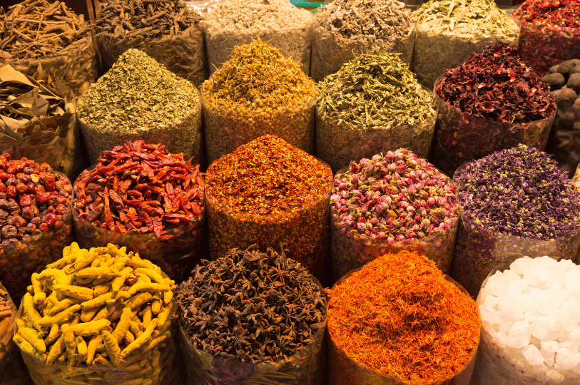 A vibrant, close-up display of spices piled high in a traditional bazaar. The foreground features bright yellow turmeric roots, star anise, and vivid red saffron, while the background shows rows of dried chili peppers, hibiscus flowers, and aromatic herbs.