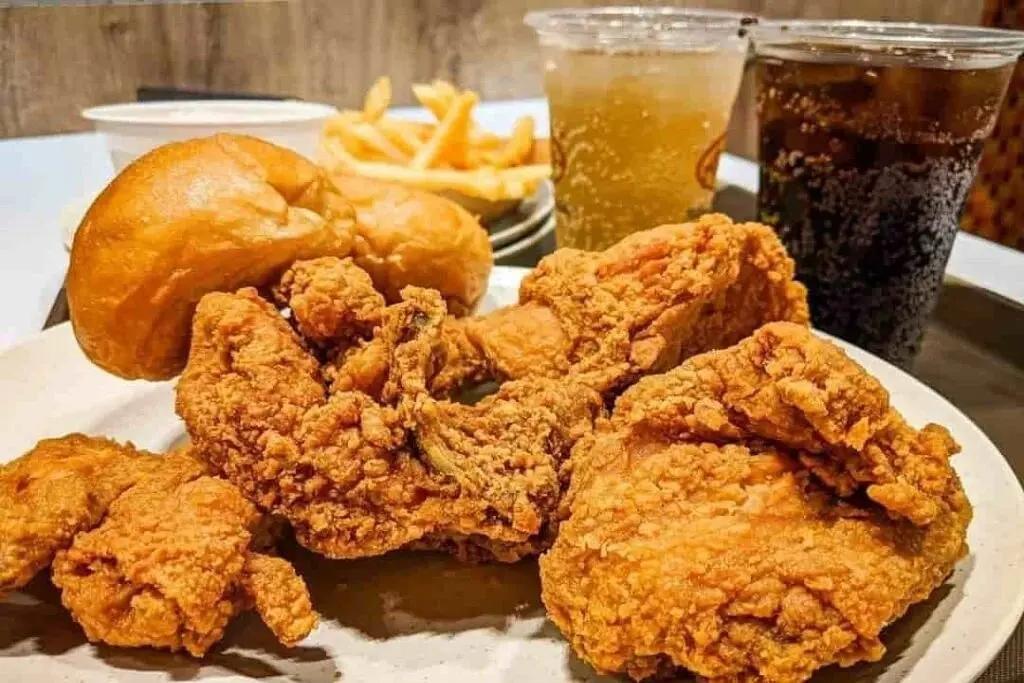 A large plate of golden, crispy fried chicken takes center stage, accompanied by two soft buns. In the background, french fries and two iced drinks complete the fast-food meal setting.