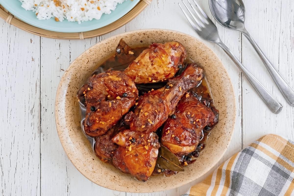 Top-down view of traditional Filipino chicken adobo served in a bowl with rich soy-vinegar sauce, bay leaves, peppercorns, steamed rice, and dining utensils.