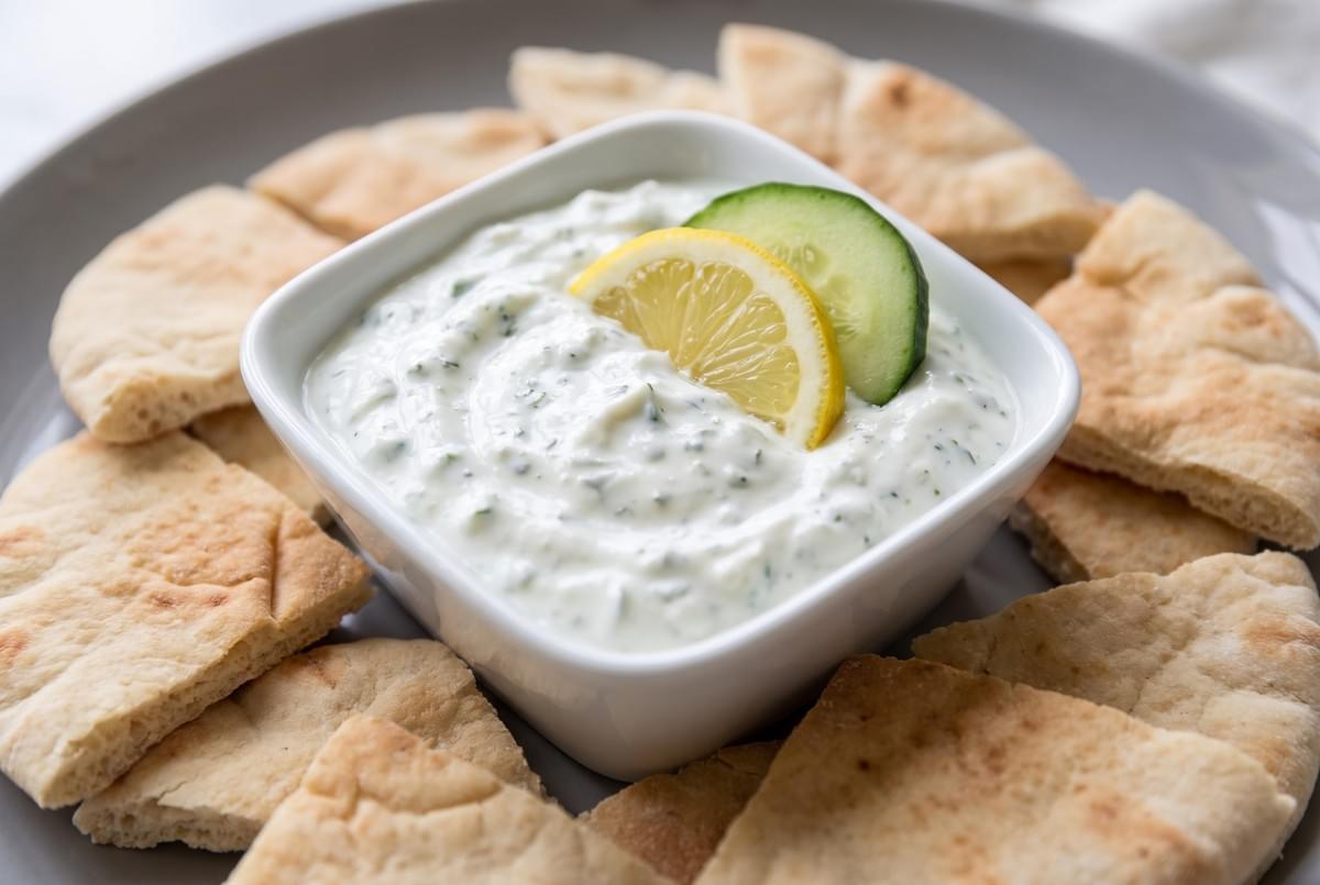 Small bowl of creamy tzatziki dip garnished with lemon and cucumber slices, surrounded by pita bread triangles.