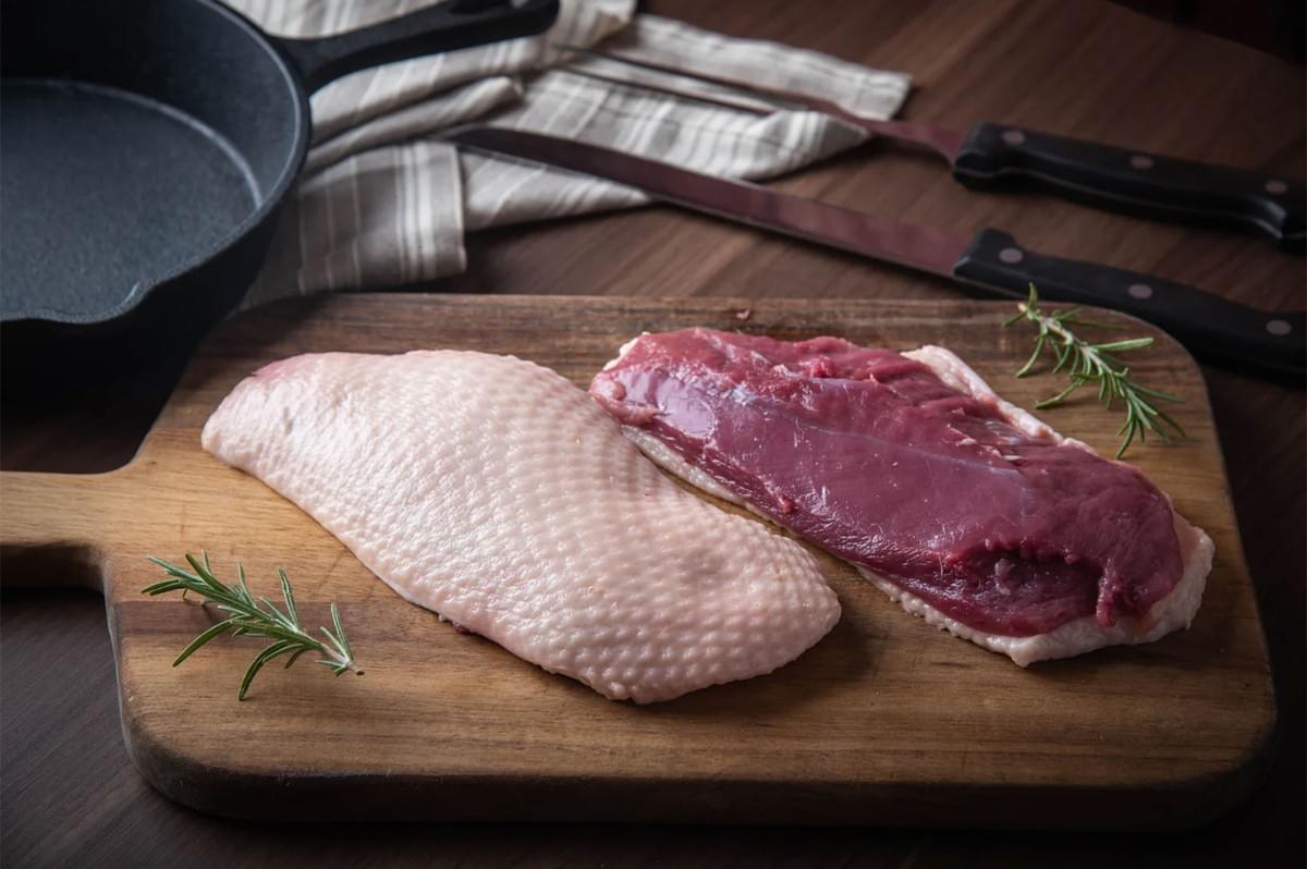 Two raw duck breasts resting on a wooden cutting board, one showing the textured skin side and the other showing the deep red meat side, garnished with fresh rosemary sprigs near a cast iron skillet.