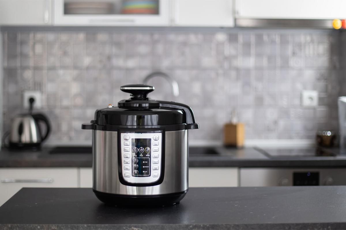 A stainless steel electric multi-cooker or Instant Pot sitting on a black kitchen counter, with a tiled kitchen backsplash blurred in the background.