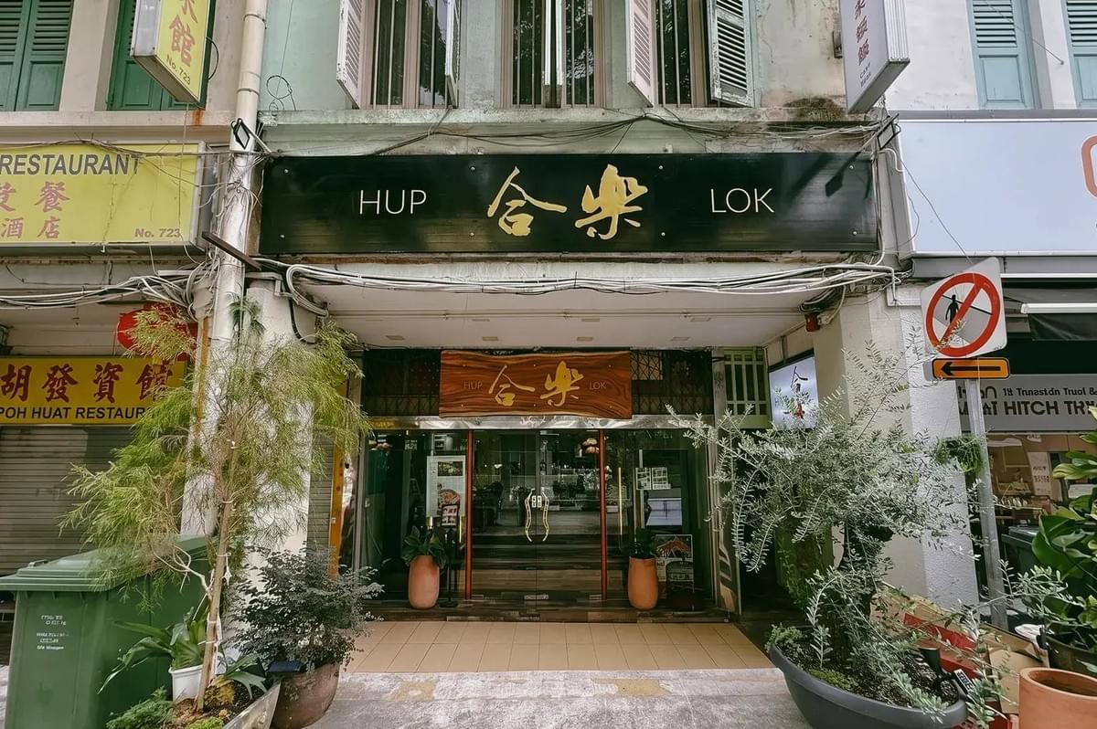 The storefront of "HUP LOK" restaurant features a large black sign with gold Chinese and English lettering above a glass entrance. Potted green plants and small trees frame the tiled walkway leading up to the shop front.