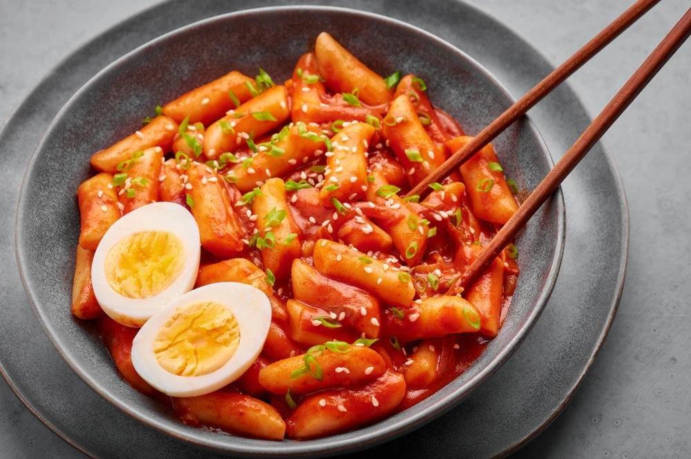 A bowl of chewy Korean rice cakes is smothered in a vibrant, spicy red gochujang sauce and garnished with sesame seeds and scallions. Two halves of a hard-boiled egg rest on the side, accompanied by a pair of wooden chopsticks ready for a meal.