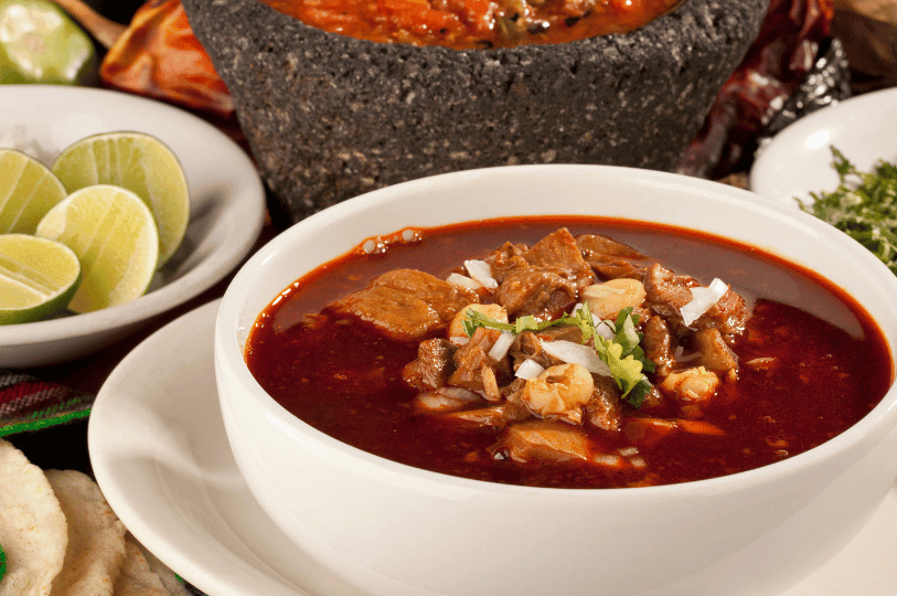 A close-up of Pozole Rojo in a white bowl with a dark, concentrated red chili broth, topped with fresh cilantro and diced onions, with a volcanic rock molcajete in the background.