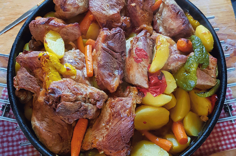 A hearty pan of Croatian meat Peka featuring thick cuts of roasted pork or veal, surrounded by whole baby carrots, yellow potatoes, and roasted green and yellow bell peppers.