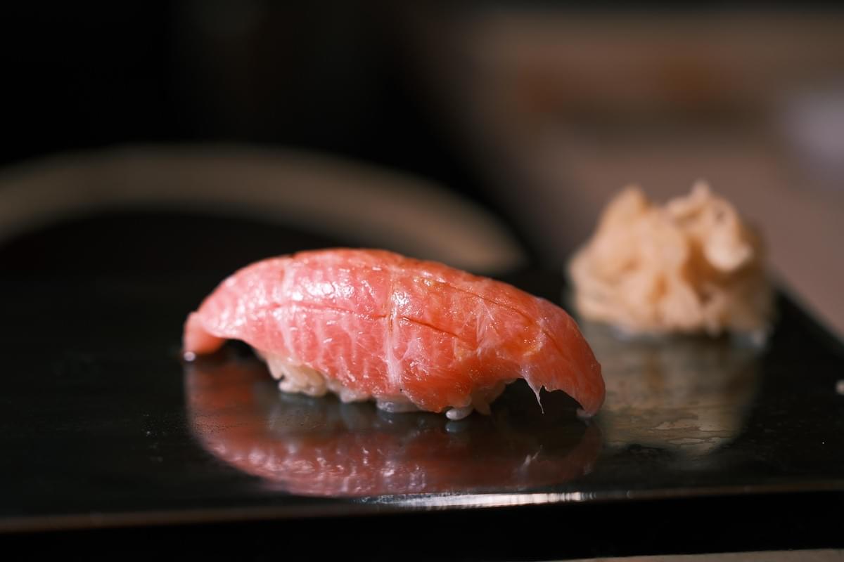 A fatty piece of tuna nigiri, showing delicate marbling and a glossy sheen, is presented on a dark, reflective surface. The composition features a tight focus on the fish, with a side of ginger resting in the soft-focus background.