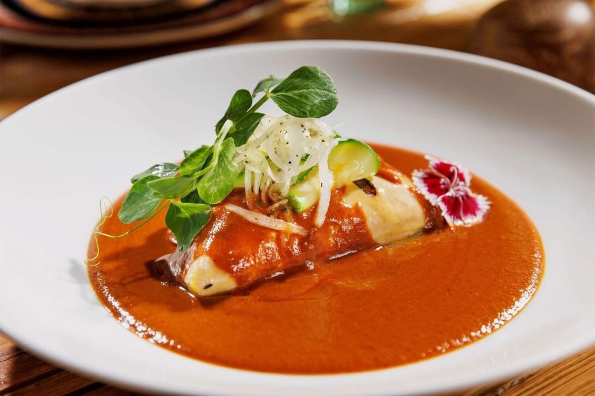 Close-up of a refined, white-plated dish (possibly a modern take on an enchilada or enmolada) featuring chicken or fish covered in red mole sauce, garnished with microgreens, shredded onion, and a delicate edible flower.