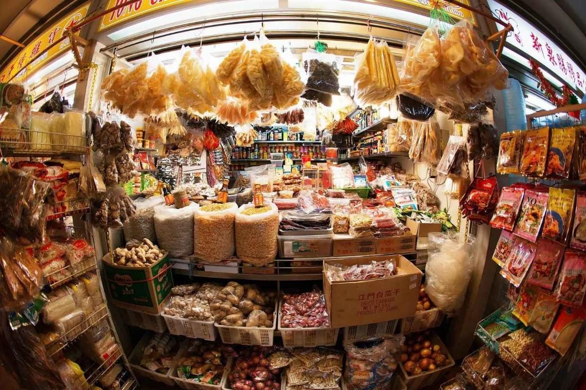 This image showcases a densely packed dry goods stall at a market, filled with a wide variety of preserved ingredients and pantry staples. Bags of dried seafood and snacks hang from the ceiling, while the lower shelves and floor are lined with crates of onions, ginger, garlic, and large sacks of grains.