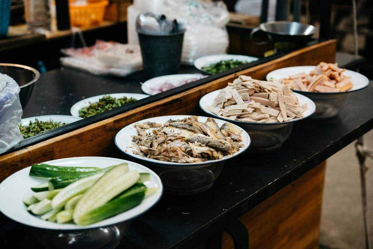 A variety of fresh ingredients, including sliced cucumbers, fried fish, and cold cuts, are neatly arranged in white bowls on a dark counter. These toppings appear ready for assembly at a street food stall, with utensils and herbs visible in the background.