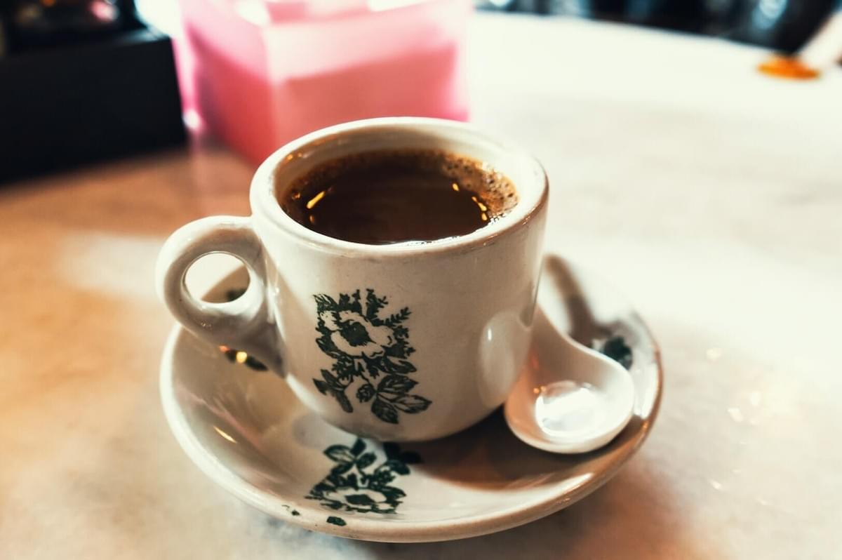 A steaming cup of coffee sits in a white cup with floral patterns, placed on a matching saucer with a spoon on a marble tabletop, evoking a cozy café ambiance.