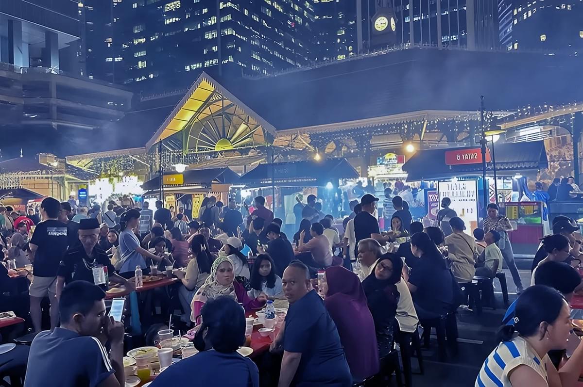 A bustling night market with packed tables of people enjoying food. Stalls glow under string lights, and skyscrapers loom above, adding urban ambiance.