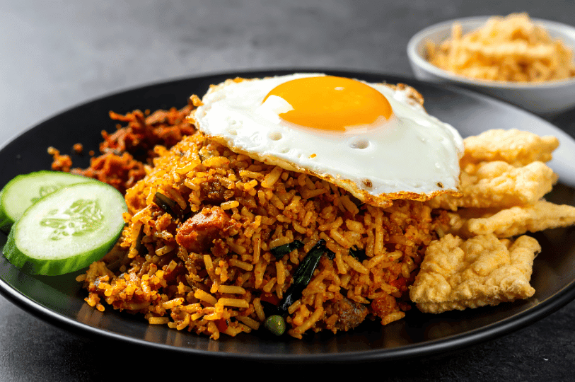 A close-up of a plate of Nasi Goreng topped with a perfectly fried sunny-side-up egg with a runny yolk, served with sliced cucumbers, crispy kerupuk (prawn crackers), and spicy sambal on a dark plate.