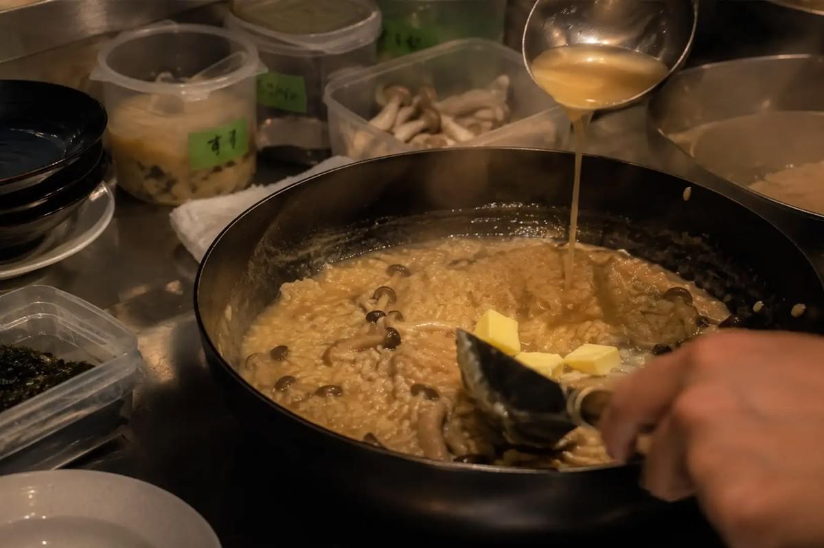 A warm, action-oriented shot inside a kitchen showing a chef pouring a ladleful of broth into a wide black pan filled with simmering rice and shimeji mushrooms, as several solid cubes of yellow butter melt into the hot, creamy mixture.