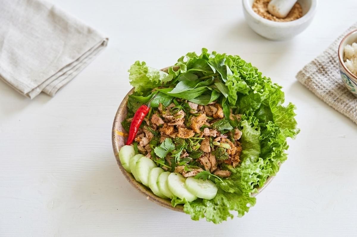 A finished bowl of Laotian Larb topped with fresh lettuce and herbs, served with cucumber slices