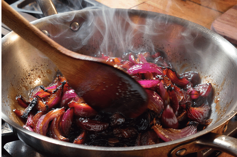 A stainless steel pan on a stovetop filled with sliced red onions beginning to soften and brown; a wooden spoon stirs the mixture as steam rises from the heat.