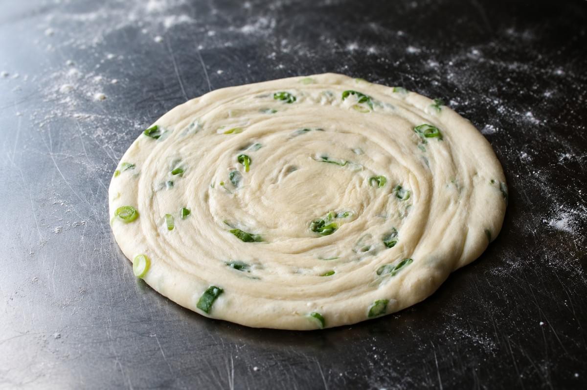 Uncooked scallion pancake dough rolled into a spiral with chopped green scallions on a floured surface.
