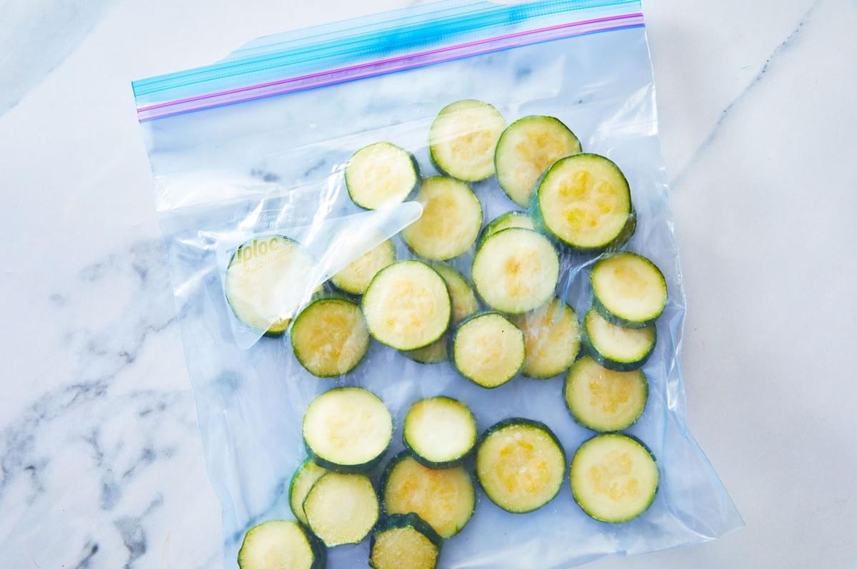 Slices of zucchini rounds frozen in a clear zip-top plastic freezer bag, resting on a white marble countertop.
