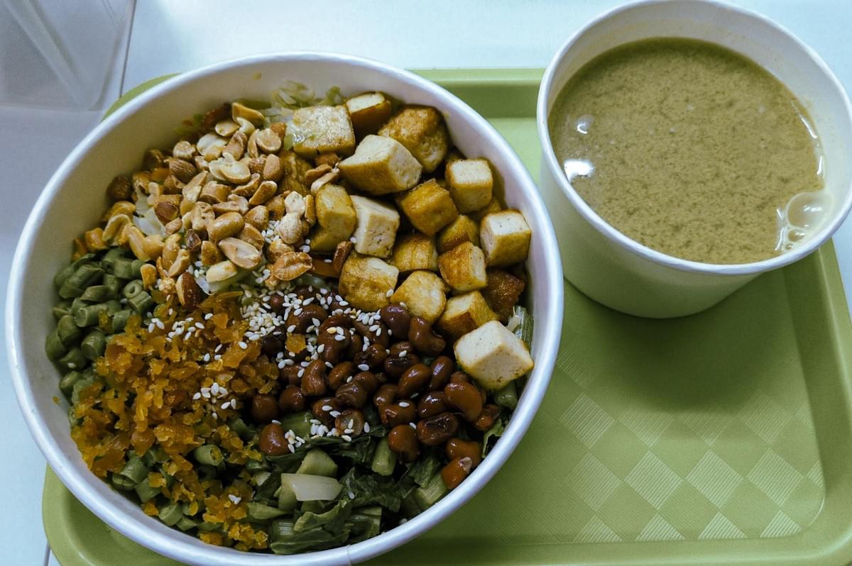 Bowl of vegetarian thunder tea rice topped with peanuts, tofu cubes, long beans, leafy greens, and preserved vegetables, served with traditional herbal soup.