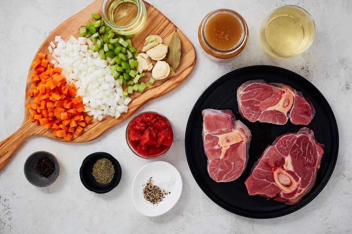 Overhead flat lay of all the raw ingredients for making Osso Buco, including raw veal shanks, diced mirepoix (carrots, celery, onion), garlic, tomatoes, stock, and wine on a white counter.