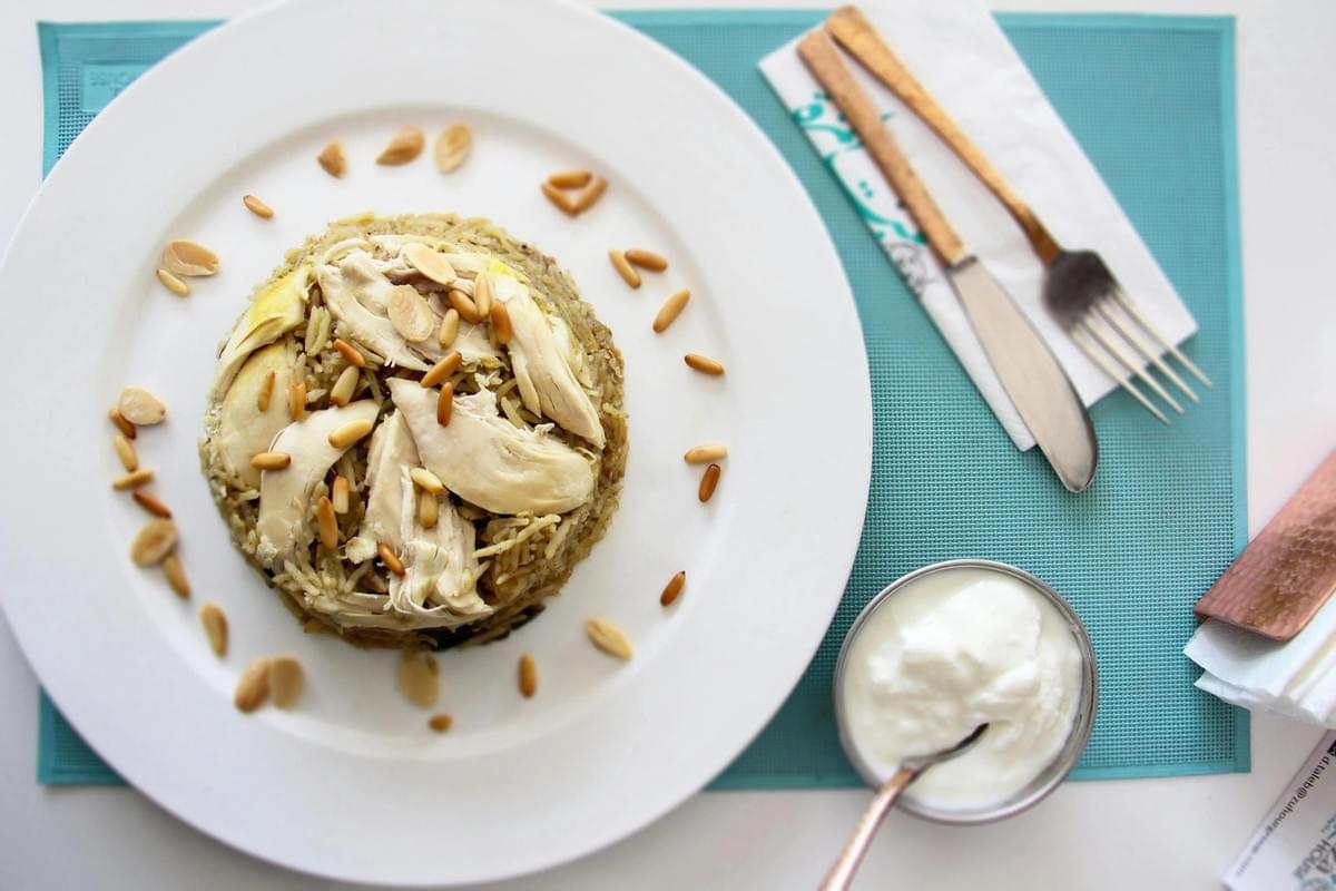 This image features a circular mound of seasoned rice topped with sliced chicken and toasted pine nuts, presented on a large white plate. Beside the main dish sits a small bowl of creamy white yogurt and a pair of gold-toned utensils resting on a white napkin.