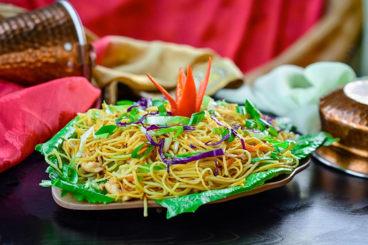 A vibrant plate of stir-fried noodles sits atop a green leaf base, colorfully garnished with shredded purple cabbage, fresh scallions, and a red chili carved into a flower. In the background, hammered copper vessels and draped red and gold fabrics create a rich, traditional setting for the meal.