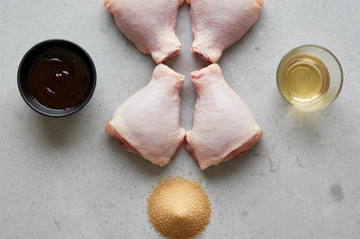 Top-down view of four raw chicken thighs arranged with bowls of dark tamarind marinade, rice vinegar, and a pile of brown sugar on a grey countertop.