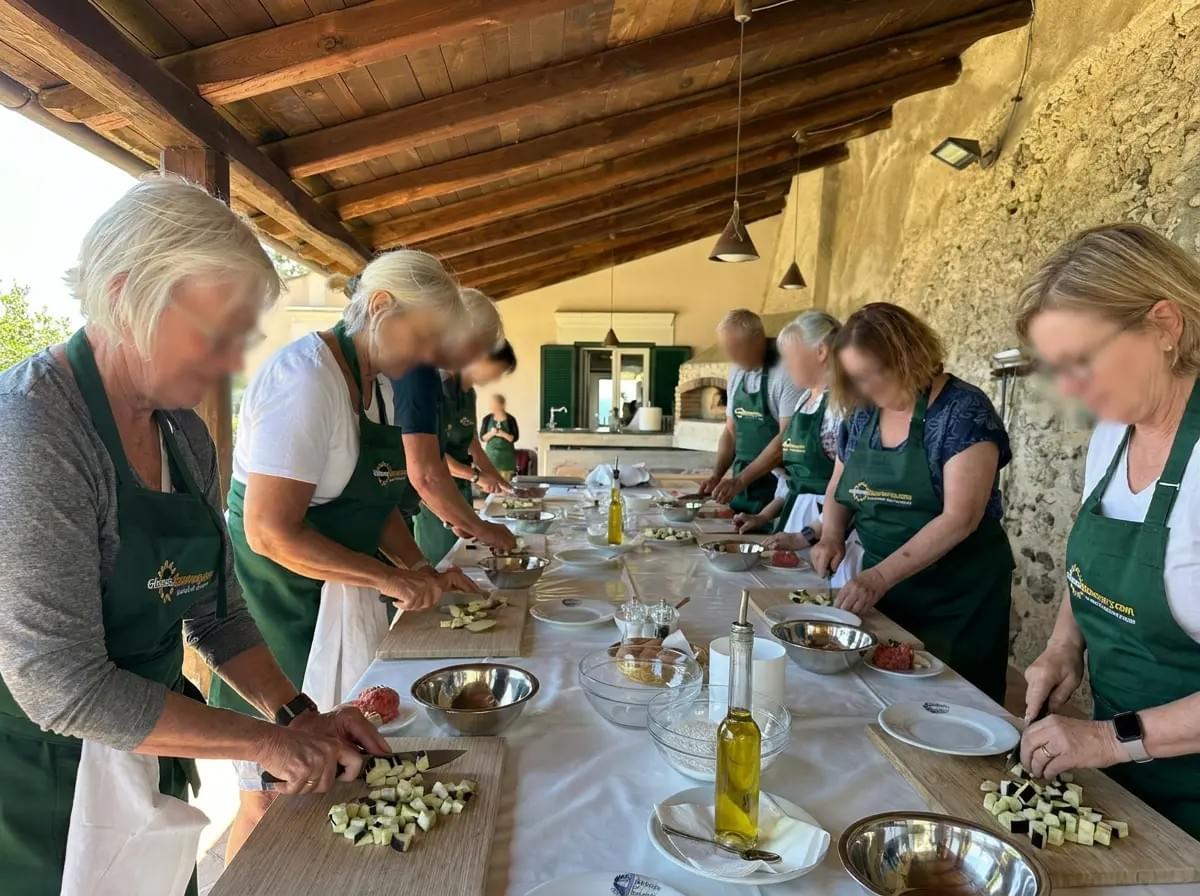 A group of people wearing green aprons stand along a long, white-clothed table under a wooden pavilion, actively chopping vegetables on wooden cutting boards during an outdoor cooking class.