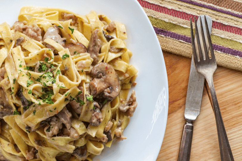 An overhead shot of a white plate loaded with creamy Russian Beef Stroganoff and mushrooms served over flat pasta, garnished with fresh chopped parsley, with silverware resting nearby.