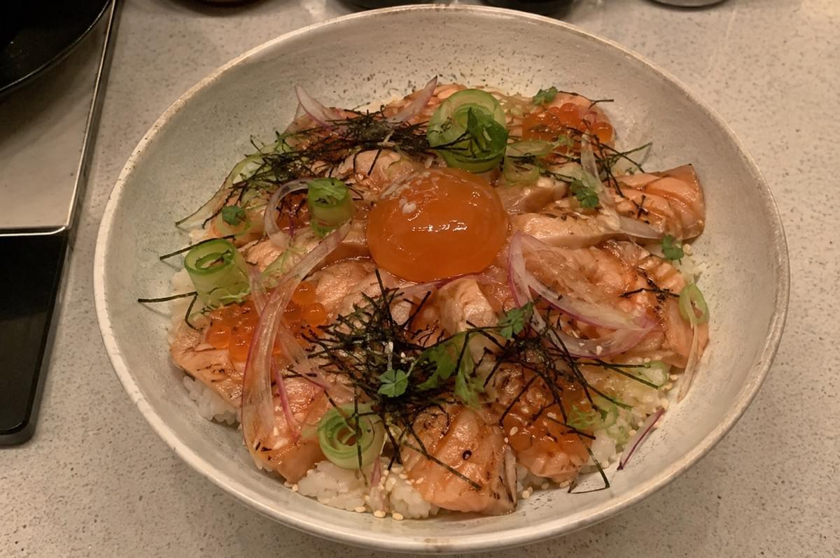 A top-down shot showcases a wide, shallow beige ceramic bowl filled with an aburi salmon chirashi bowl. Lightly seared, flame-torched salmon slices cover a bed of sushi rice, garnished with a vibrant, glossy soy-cured egg yolk centered on top. The dish is finished with clusters of glowing orange salmon ikura, finely shredded nori seaweed, thin slices of red onion and pickled myoga, curled green scallions, and small microgreens.