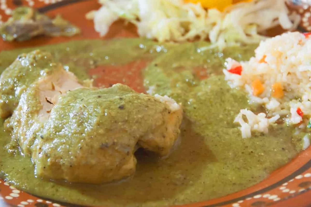 Close-up of a piece of white meat (chicken or fish) covered in a pale green Mole Verde sauce, served on a terracotta plate with white rice and shredded lettuce.