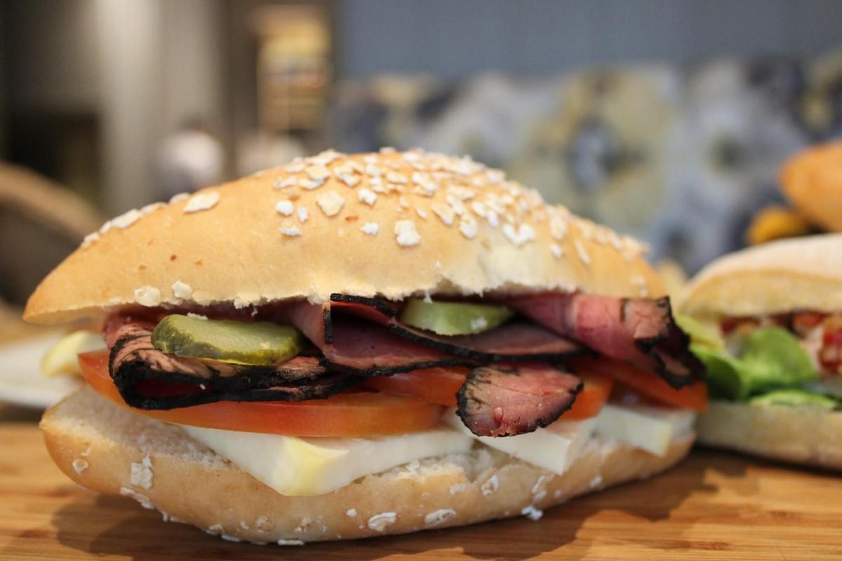 A close-up shot of a sandwich filled with thin slices of roast beef, tomato, pickles, and cheese served on an oat-topped roll. The sandwich rests on a wooden board against a softly blurred background.