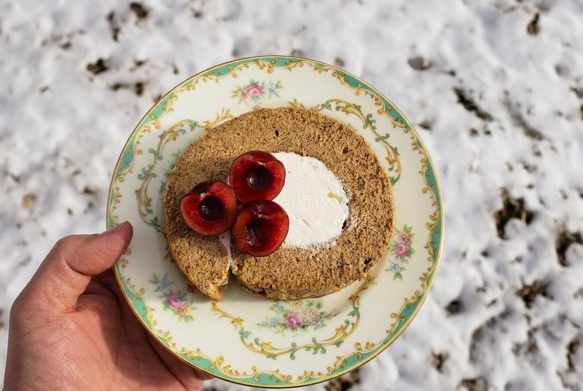 Hand holding a vintage floral plate with a slice of cream-filled roll cake topped with fresh cherry halves.