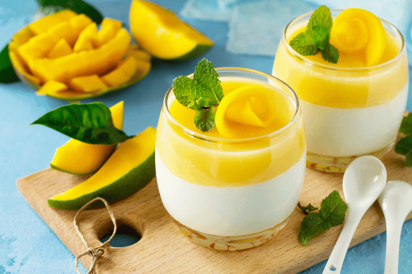 Two elegant glass tumblers featuring a thick layer of white Panna Cotta topped with bright yellow mango puree and a decorative mango swirl garnish.