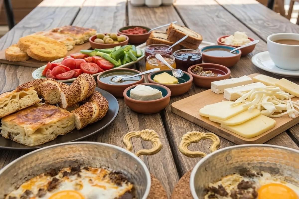 Lavish Turkish breakfast spread on a wooden table featuring simit, assorted cheeses, olives, fresh vegetables, jams, and eggs in copper pans