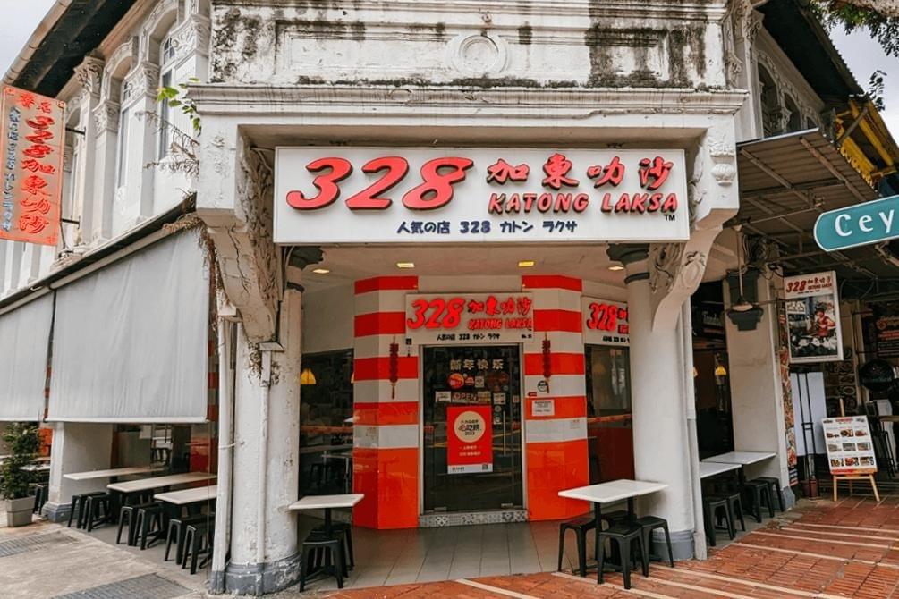 The image shows the exterior of the 328 Katong Laksa restaurant, featuring its prominent red and white signage and traditional shophouse architecture. Outdoor seating with simple white tables and black stools lines the sidewalk under an awning, inviting diners to enjoy a meal in an open-air setting.
