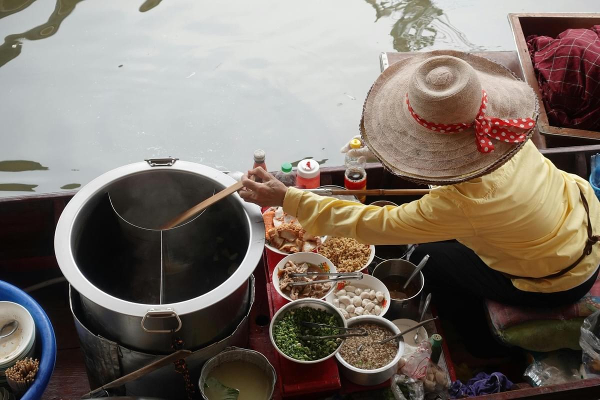 Dressed in a yellow shirt and a straw hat with a red polka-dot ribbon, a vendor tends to a large steaming pot aboard a small boat. The floating workspace is crowded with bowls of fresh ingredients, such as chopped green onions, sliced meats, and fish balls, set against the backdrop of the water.