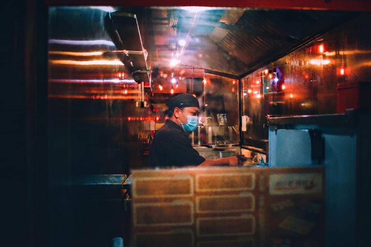 Chef wearing a face mask cooking in a small street food stall at night.