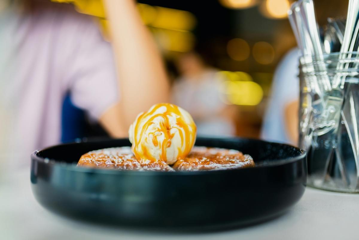 The image shows the exterior of the Tekka Centre in Singapore, a vibrant building with teal, yellow, and blue geometric accents. People are seen walking across a tiled plaza in the foreground under the shade of large, leafy trees.