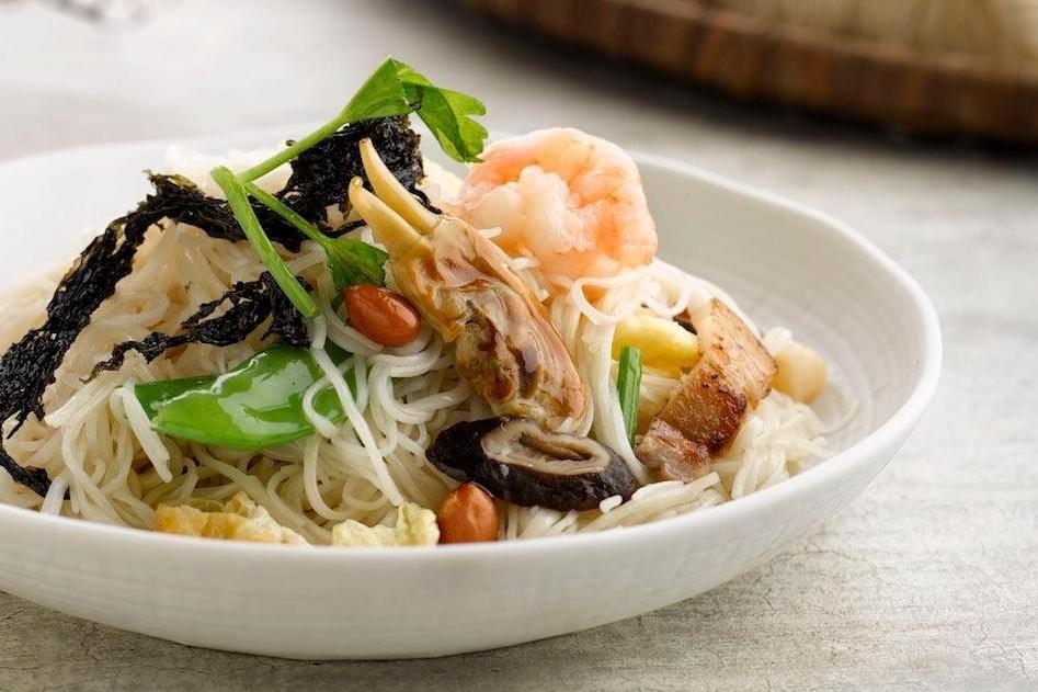 The image features a white bowl filled with thin rice noodles topped with shrimp, clams, and sliced pork. The dish is garnished with dark seaweed, green vegetables, and peanuts to create a colorful and textured meal.