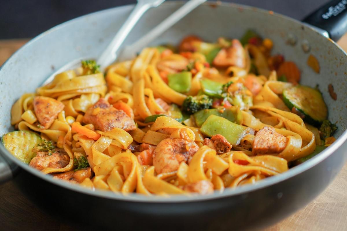 A skillet filled with cooked fettuccine or egg noodles tossed with grilled chicken pieces and mixed vegetables, including broccoli, snap peas, and zucchini. Two white utensils rest inside the pan, ready for serving.