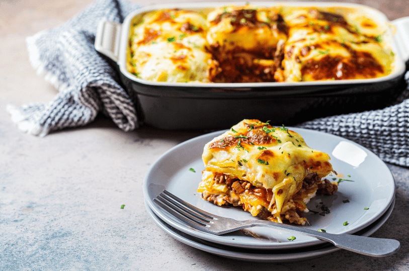 A golden slice of Greek moussaka layered with roasted eggplant, spiced ground meat, and creamy béchamel, served on a white plate with lemon wedges, parsley, and mint-topped yogurt sauce—evoking Mediterranean warmth and layered comfort.
