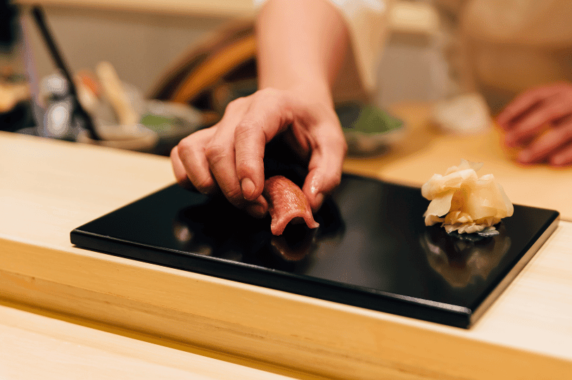 A sushi chef places a piece of tuna nigiri beside pickled ginger on a black plate, highlighting the seasonal finesse and meticulous presentation found in Singapore’s authentic Japanese restaurants.