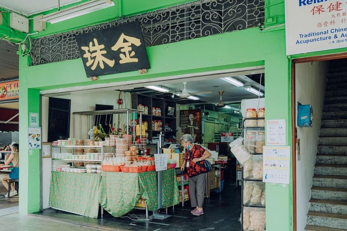 This image displays the vibrant green storefront of Kim Choo Kueh Chang, a traditional shop filled with various snacks and containers of food. An elderly woman stands near the counter inside the open-air entrance, which is marked by a prominent black sign with gold Chinese characters.