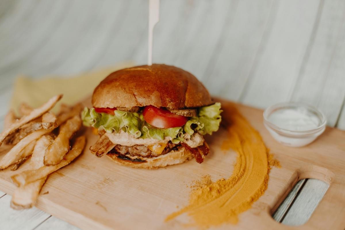 A gourmet burger topped with lettuce, tomato, and bacon sits on a wooden board alongside a pile of hand-cut fries. The presentation is accented by a decorative sweep of yellow spice and a small glass bowl of white dipping sauce.