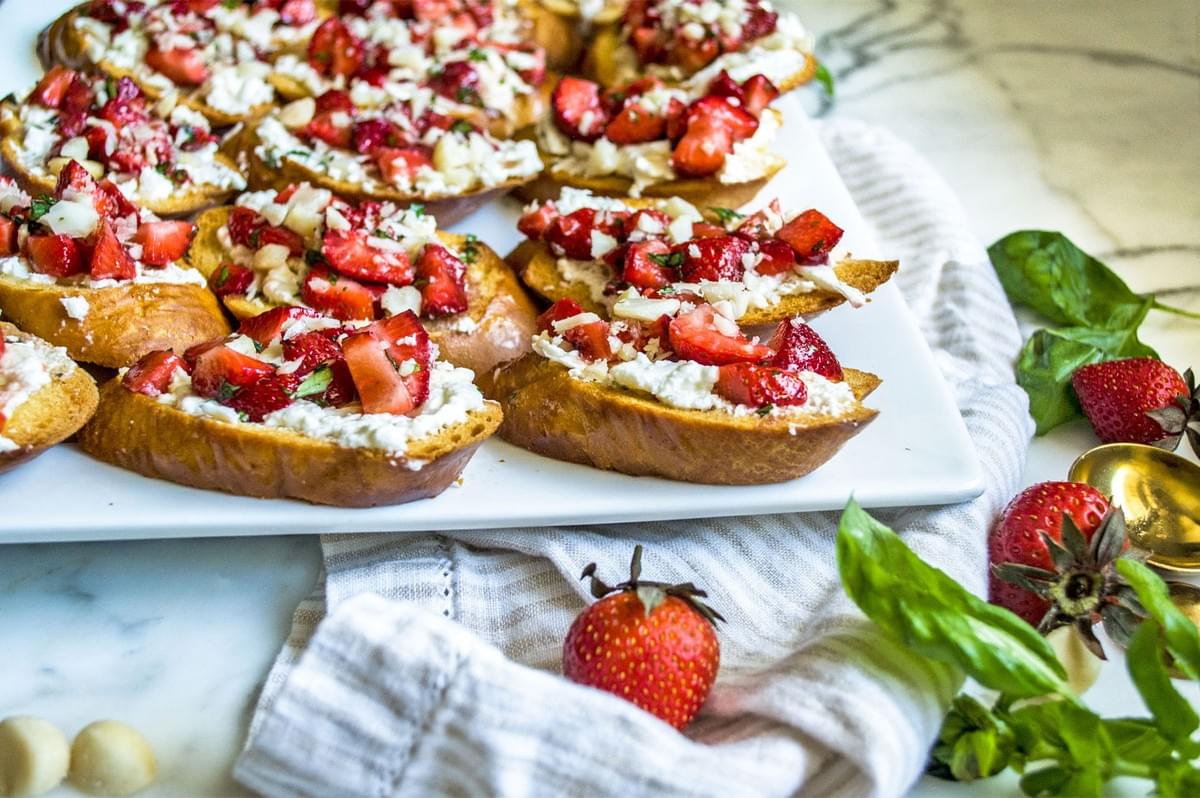 A platter of strawberry and ricotta bruschetta on sliced toasted bread, topped with a balsamic glaze and herbs, with a fresh strawberry in the foreground.
