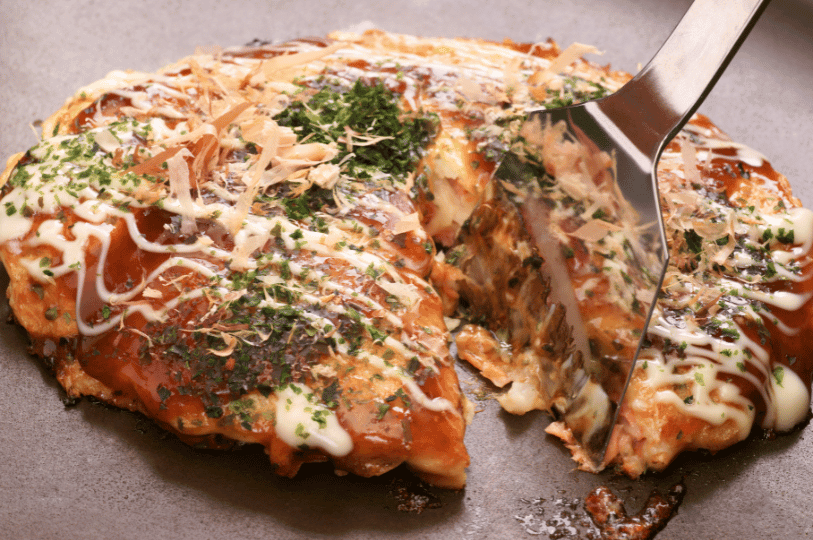 A golden okonomiyaki sizzling on a hot griddle, layered with creamy mayo, tangy sauce, aonori, and dancing bonito flakes—its crispy edge lifted by a spatula to reveal the hearty, savory interior of Japan’s beloved street food.