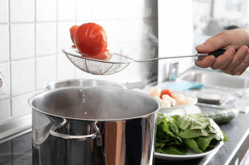 A whole tomato being lifted out of a steaming pot of boiling water with a wire skimmer, demonstrating the blanching technique used for peeling.