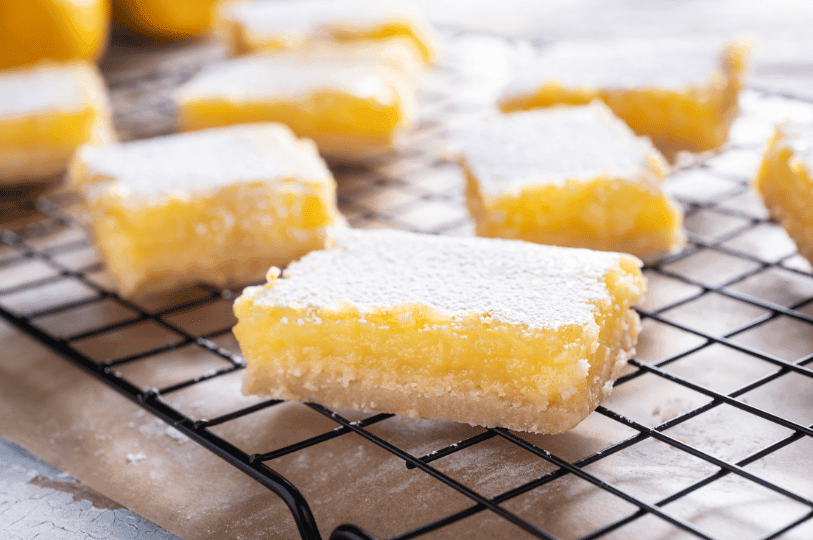 A low-angle shot of lemon bars cooling on a black wire rack. The focus is on a single bar in the foreground, showing the porous texture of the powdered sugar topping and the distinct layers of tart curd and golden crust.